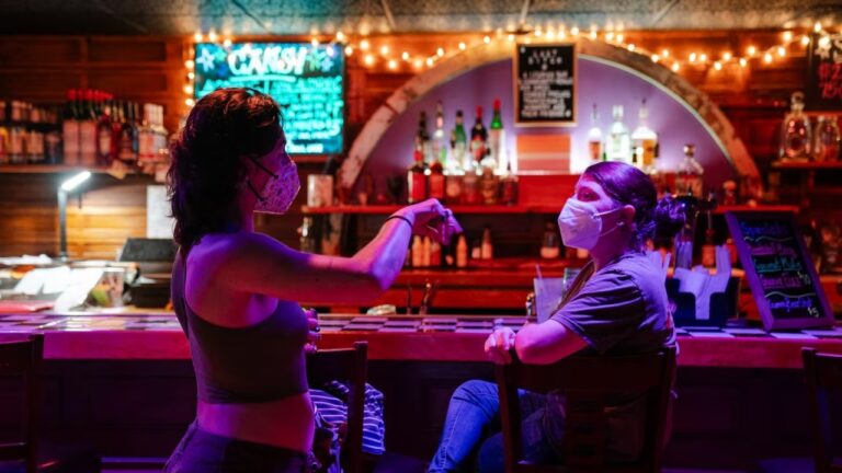 Twee vrouwen met mondkapjes in gesprek aan de bar van Last Ditch, in rood en paars licht met lichtslingers en flessen op de achtergrond.