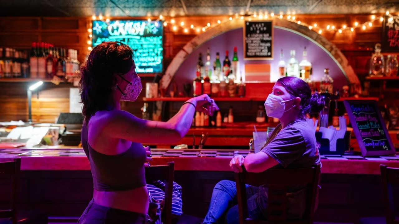Twee vrouwen met mondkapjes in gesprek aan de bar van Last Ditch, in rood en paars licht met lichtslingers en flessen op de achtergrond.